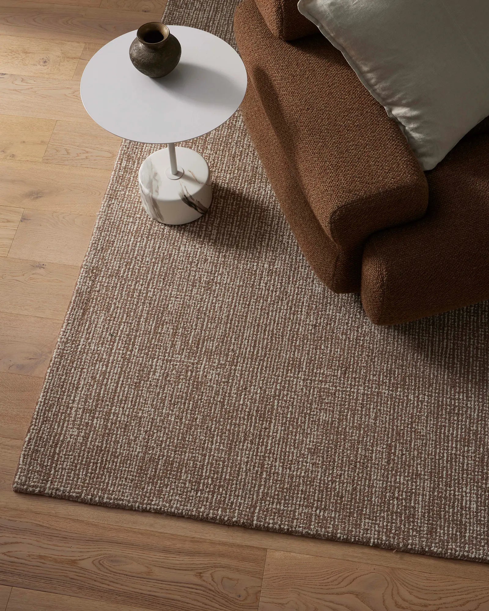 Brown textured rug on a wooden floor with a brown sofa and white side table.