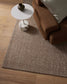 Brown textured rug on a wooden floor with a brown sofa and white side table.