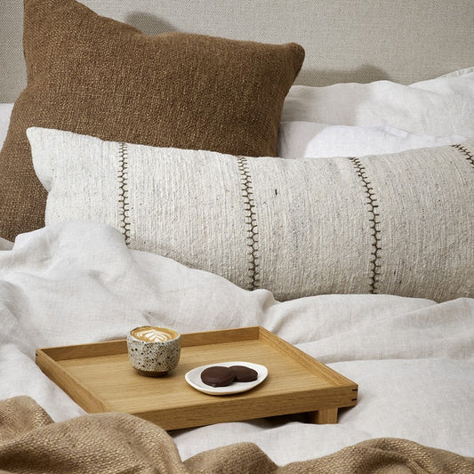 Kalahari lumbar cushion in cream with khaki embroidery on a bed. Behind is a square cocoa cushion and in front is a small bamboo tray with a coffee and plate of chocolate biscuits. The bed is in white linen with a glimpse of a begie throw