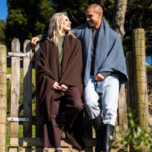 A man and a woman standing outdoors next to a wooden fence, both wrapped in thick knit blankets. The woman wears a dark brown ribbed blanket and matching brown pants, while the man wears a blue-gray ribbed blanket with light blue jeans.