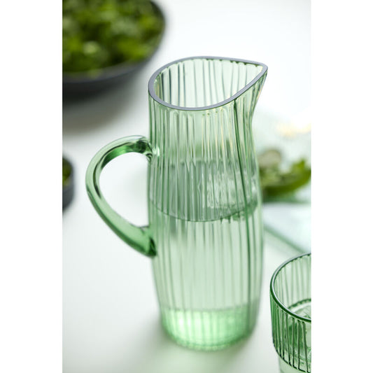 Green glass pitcher on a white surface with blurred greenery in the background