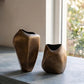 Two abstract sculptural ceramic vases in a warm metallic bronze finish, standing on a gray stone countertop next to a window.