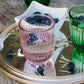 A pink ribbed glass tumbler with a red rim, partially filled with water and blueberries, displayed on a mirrored tray alongside a green glass and blueberries.