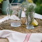 Clear glass pitcher and cups on a woven mat with a lemon and herbs, outdoor setting