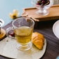 Clear glass mug with green tea on a decorative plate with a croissant and cherry, on a wooden table.