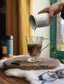 Person pouring coffee from a white mug into a glass mug on a wooden coaster.