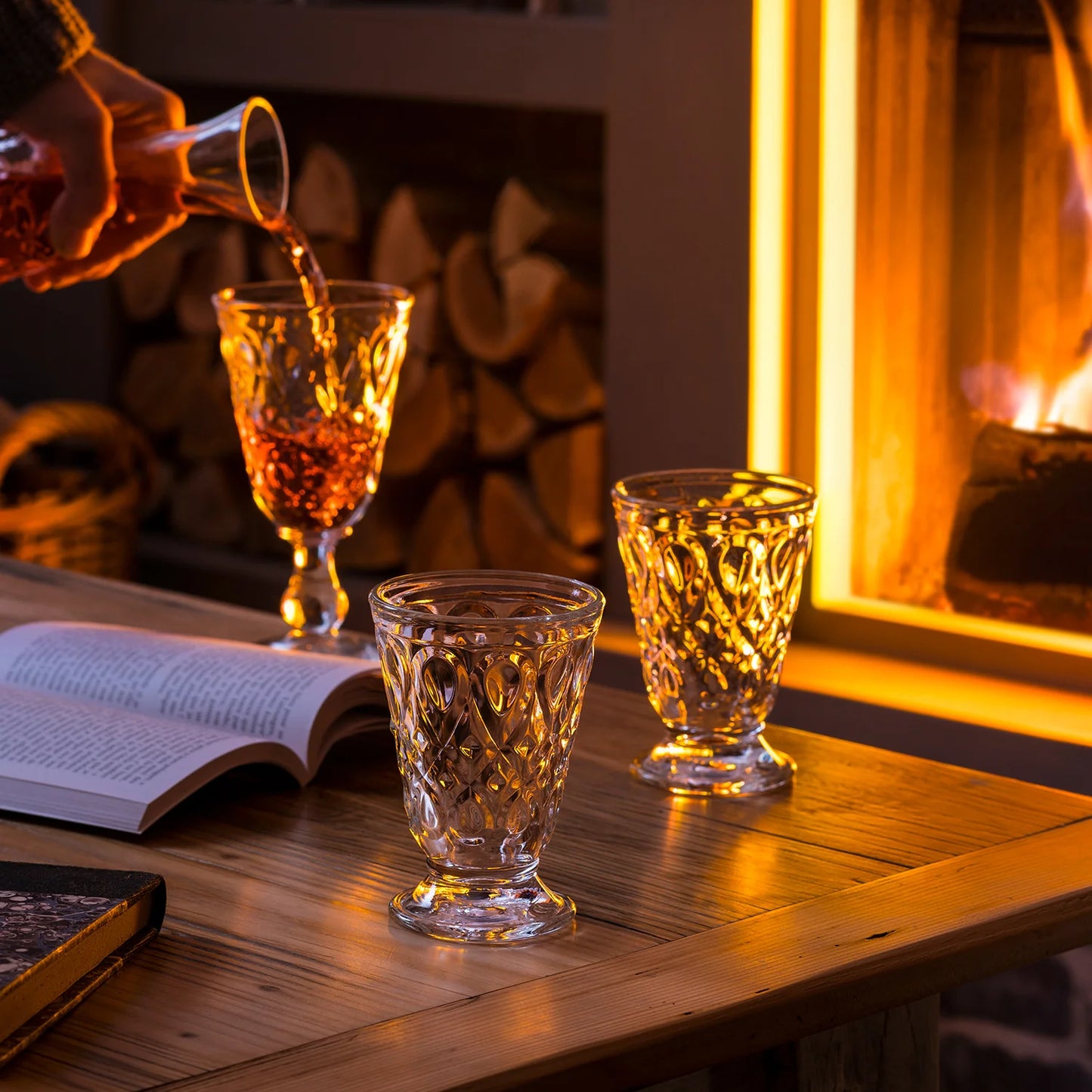 Person pouring a drink into a glass on a wooden table with a fireplace in the background