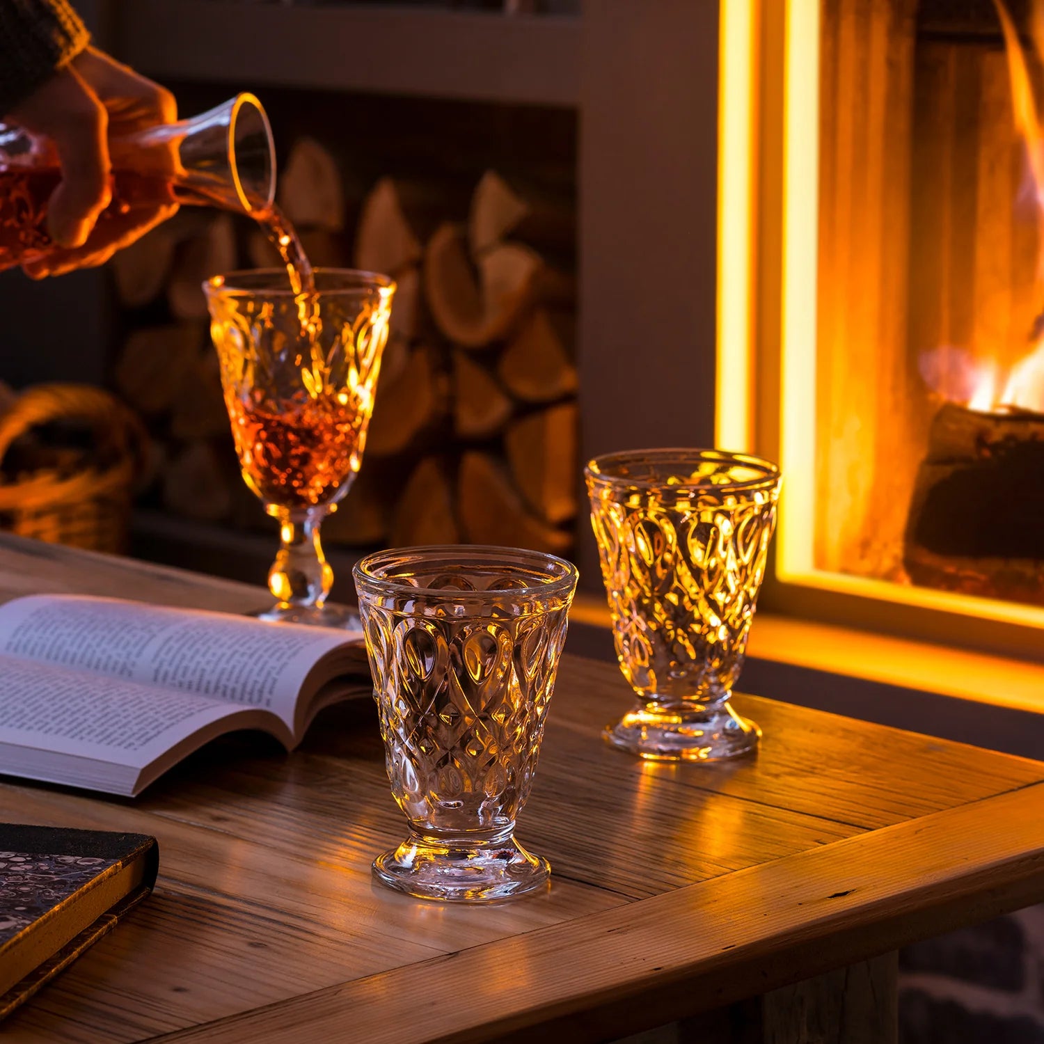 Person pouring a drink into a glass on a wooden table with a fireplace in the background
