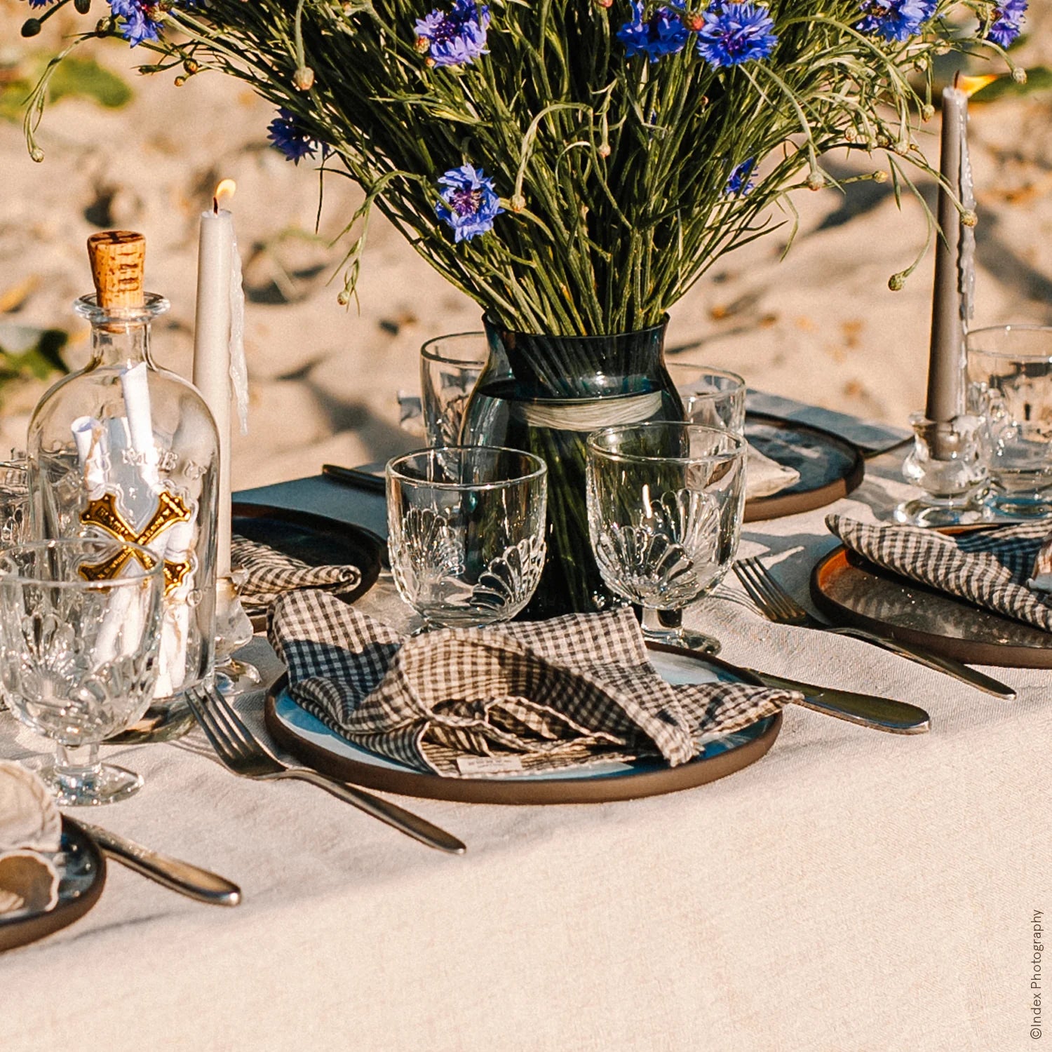Table setting with checkered napkins, glasses, and a vase of flowers on a sandy background