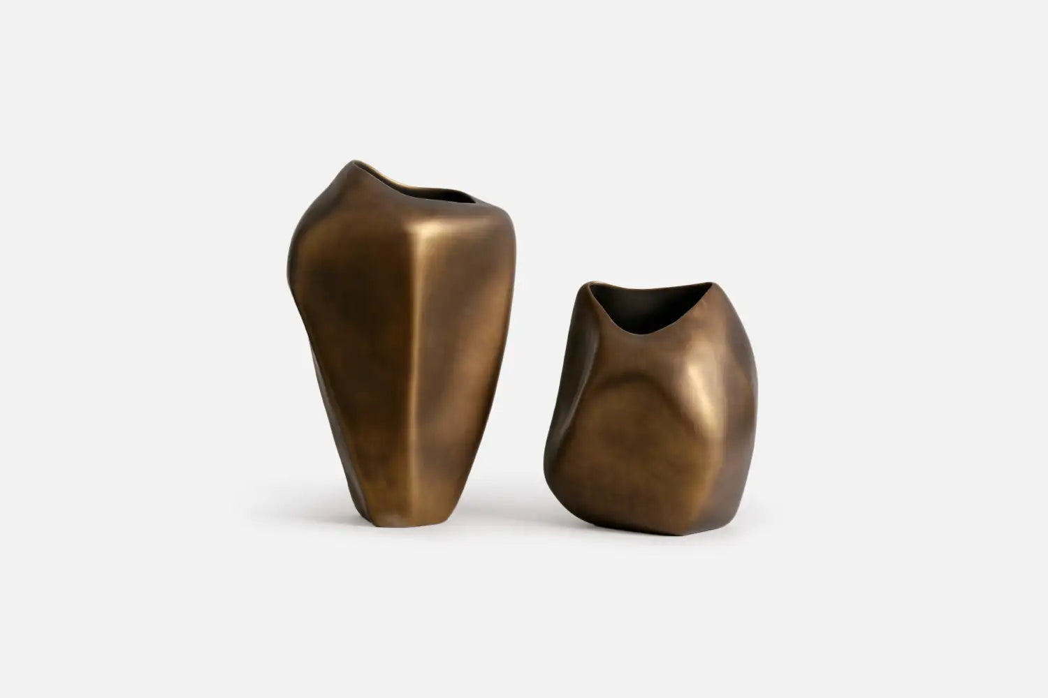 Two abstract sculptural vases in a brushed bronze finish, standing on a light gray studio background.