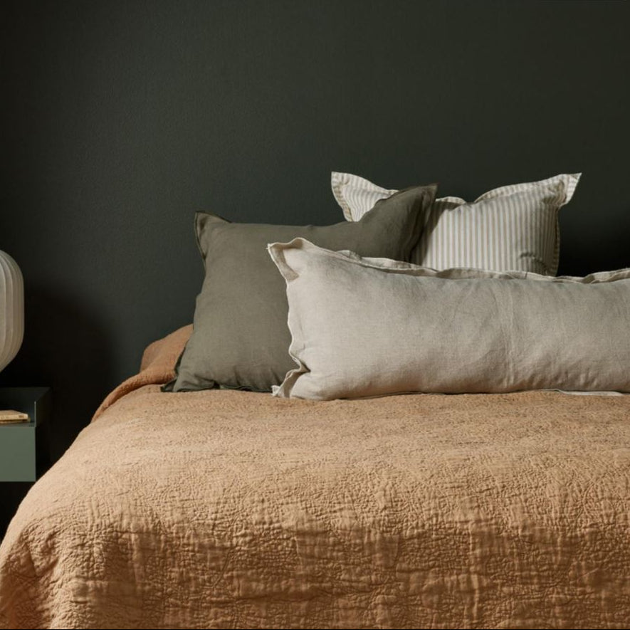 Bed with brown bedding and gray pillows against a dark wall, with a nightstand and lamp.