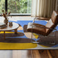 abstract rug with organic shapes in yellow, blue and brown with a modern leather chair on it and a round wooden coffee table with white decor pieces.