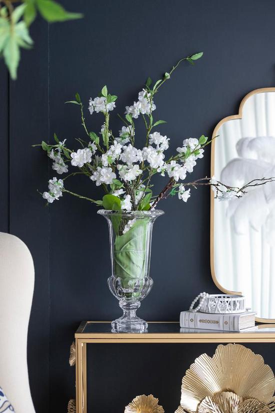 White cherry blossom branches arranged in a clear glass pedestal vase on a console table against a dark navy wall, with a gold-framed mirror and decorative objects nearby.