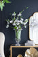 White cherry blossom branches arranged in a clear glass pedestal vase on a console table against a dark navy wall, with a gold-framed mirror and decorative objects nearby.