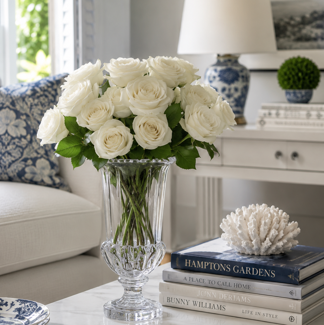 A bouquet of white roses in a clear glass vase on a table, styled in a bright living room next to a stack of books and a coral shell decor piece.