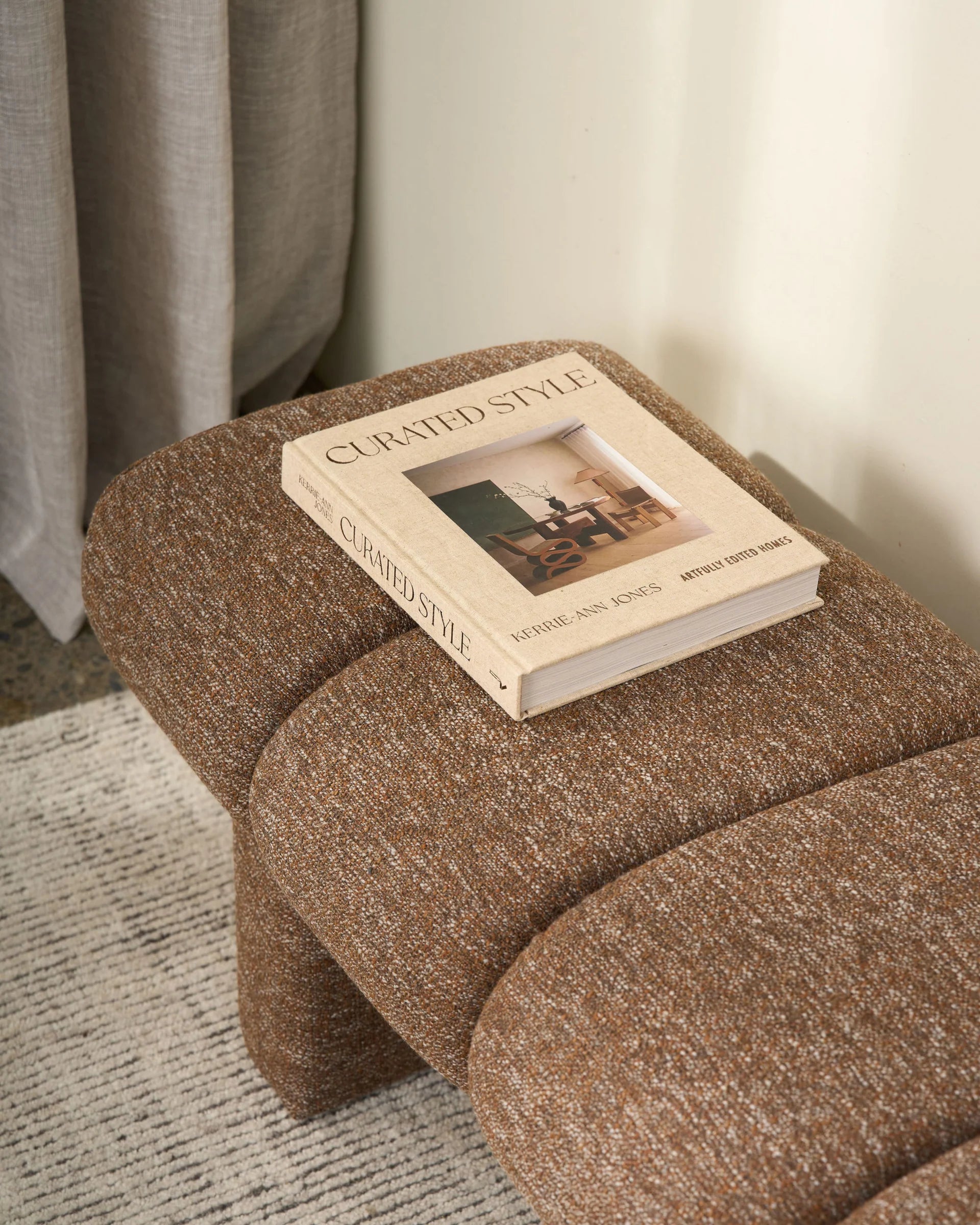Brown textured ottoman with a book titled 'Curated Style' on top, against a neutral background.