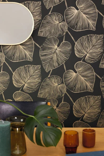 Black wallpaper with a beige leaf pattern, styled in a room setting with a wooden table, amber glass cups, a green plant, and a partial oval mirror.