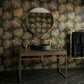 A luxury bathroom scene featuring a dark wood console table with a black marble top, a black vessel sink, and a gold faucet, set against a black wallpaper with gold leaf patterns. A round gold-framed mirror is mounted on the wall, and a gold towel rack with a black towel is visible on the right.