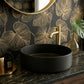 A matte black round vessel sink set into a dark marble countertop, paired with a brushed gold faucet. The background features a matching black and gold botanical wallpaper, with a gold-framed mirror and a green monstera leaf in a glass vase.