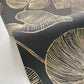 Black wallpaper with a gold botanical leaf pattern, shown partially unrolled over a white surface.