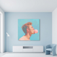 A square canvas wall art print depicting a man blowing pink bubblegum, displayed in a modern room setting on a light blue wall above a white console table.