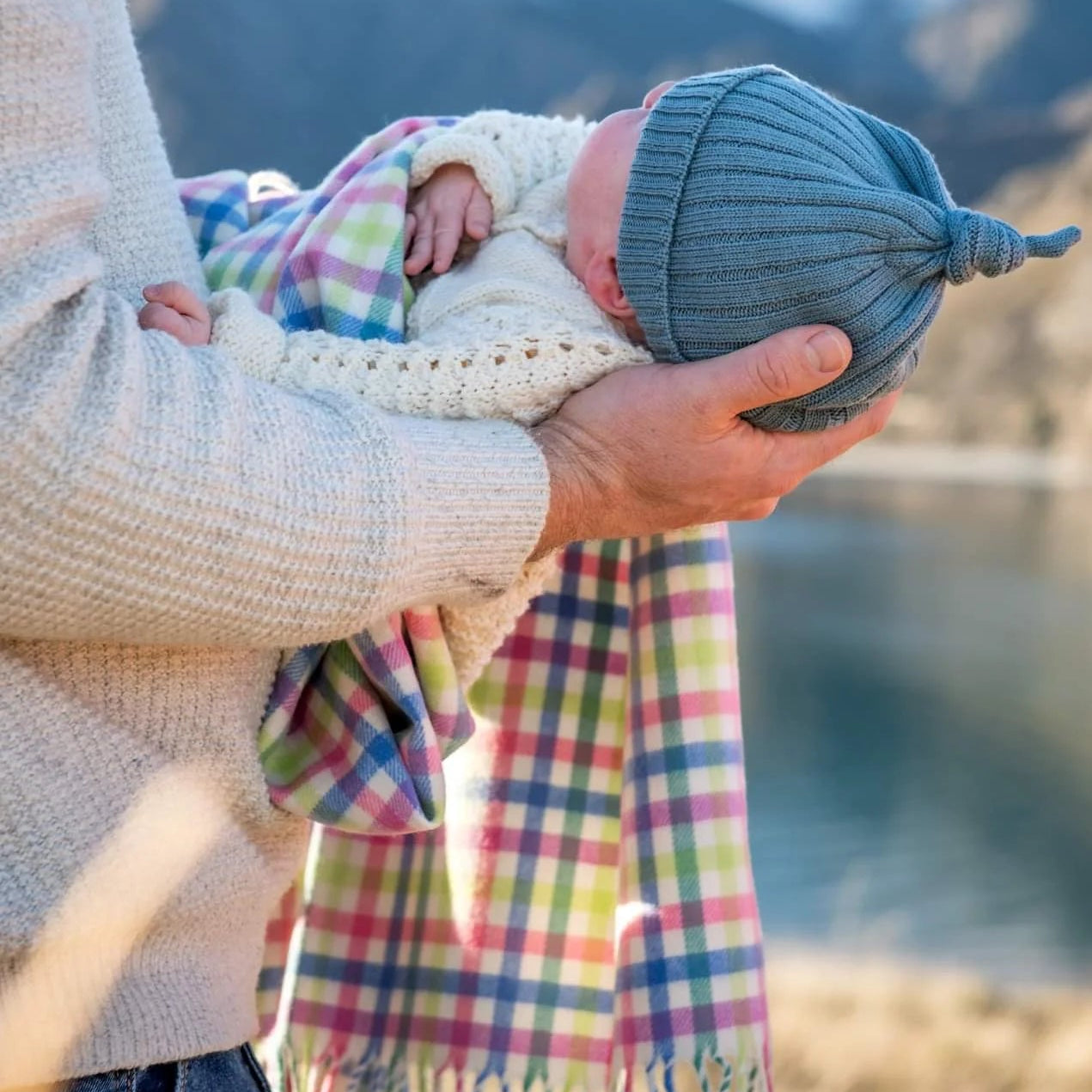 Baby in blue knitted beanie being held by a man and wrapped in a multi check merino baby blanket
