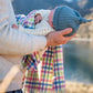 Baby in blue knitted beanie being held by a man and wrapped in a multi check merino baby blanket
