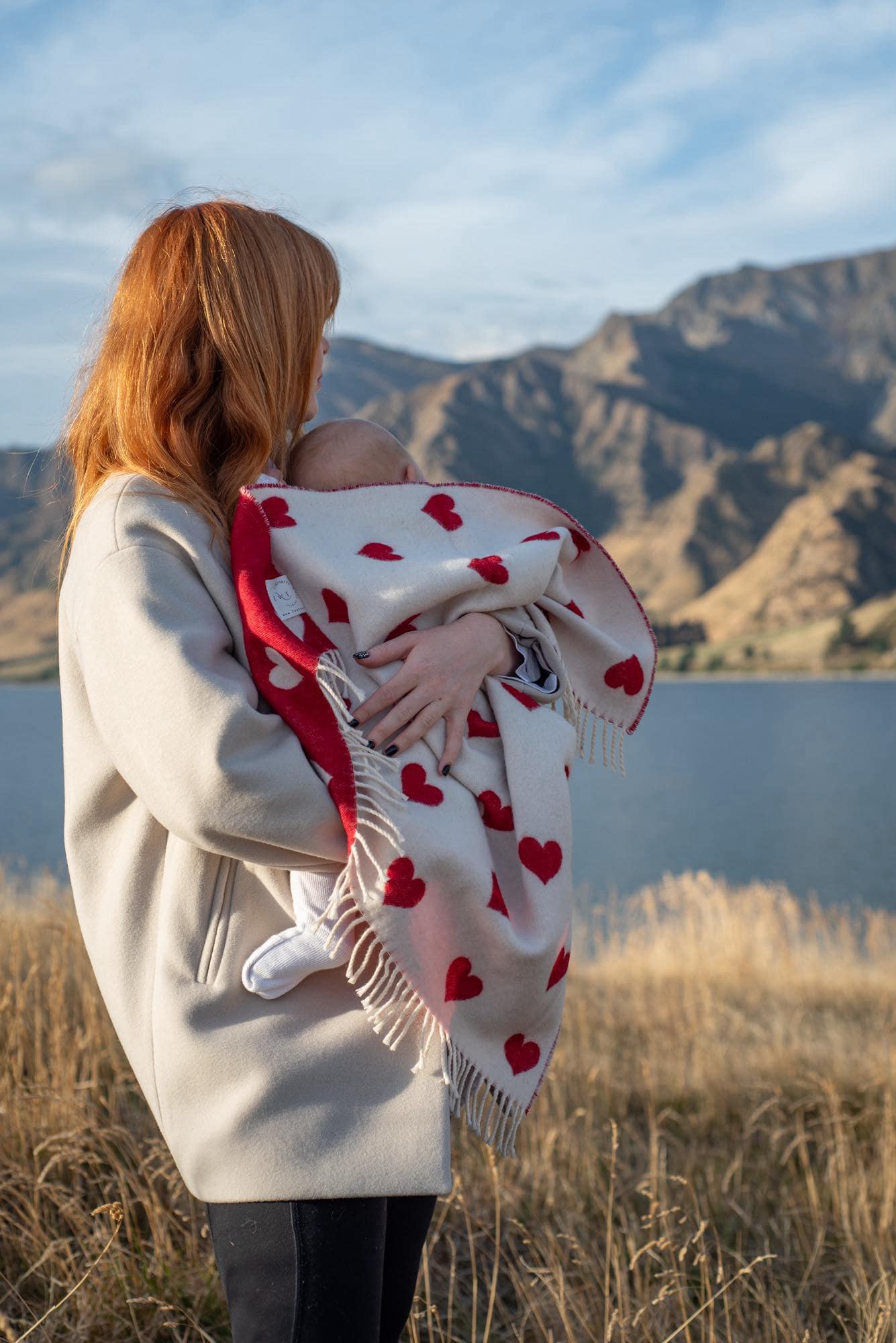 baby wrapped in a red and white hear merino blanket being held by a red haired lady mountains and lakes in the background