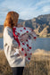 baby wrapped in a red and white hear merino blanket being held by a red haired lady mountains and lakes in the background