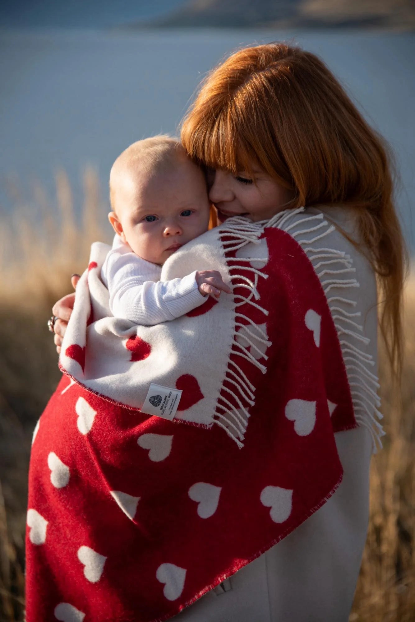 baby wrapped in a red and white hear merino blanket being held by a red haired lady, lake in background