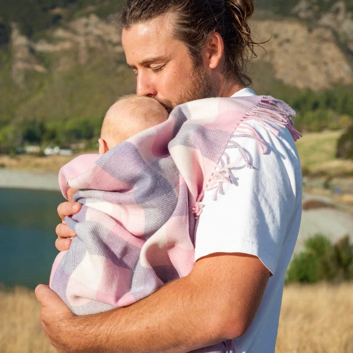 Father in white shirt holding a new born wrapped in a pink and white checked merino blanket with lake in background