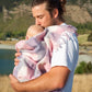 Father in white shirt holding a new born wrapped in a pink and white checked merino blanket with lake in background