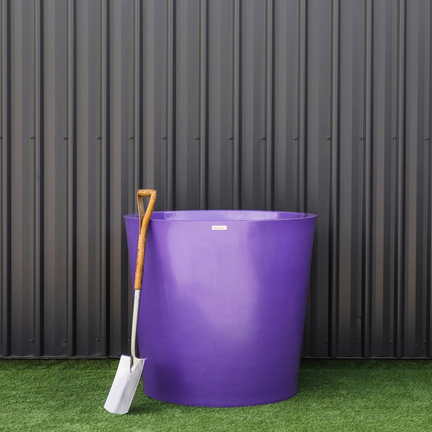 Purple plastic planter with a shovel on grass against a metallic wall