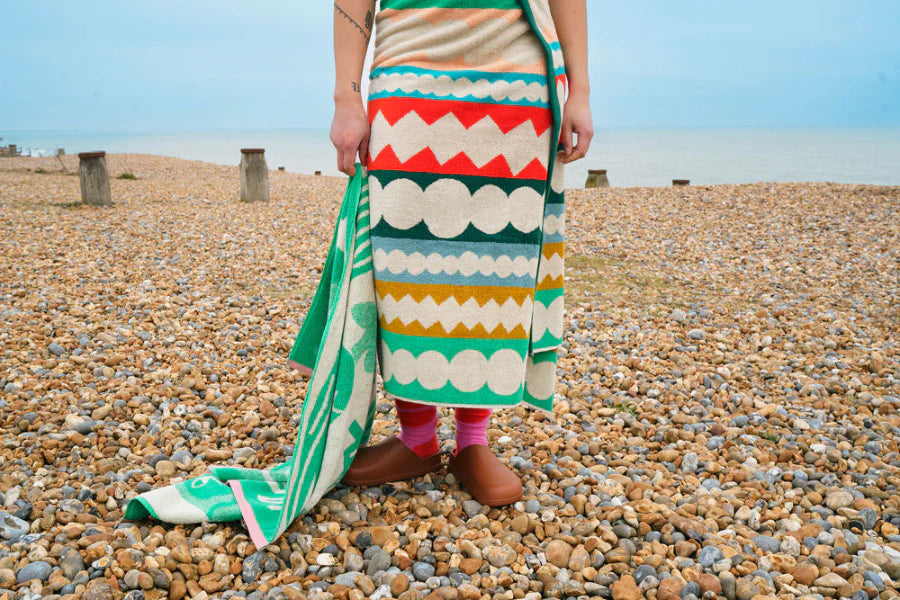 Person standing on a pebbly beach wearing a colorful patterned towel.