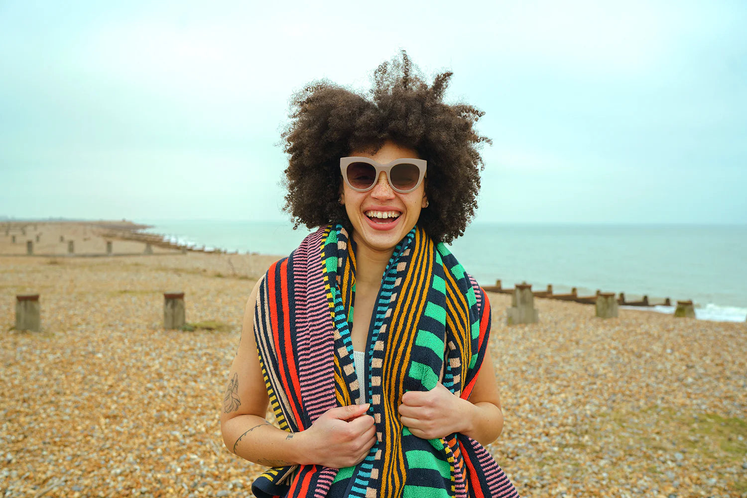 Woman with an afro wearing sunglasses and a colorful towel on a beach.