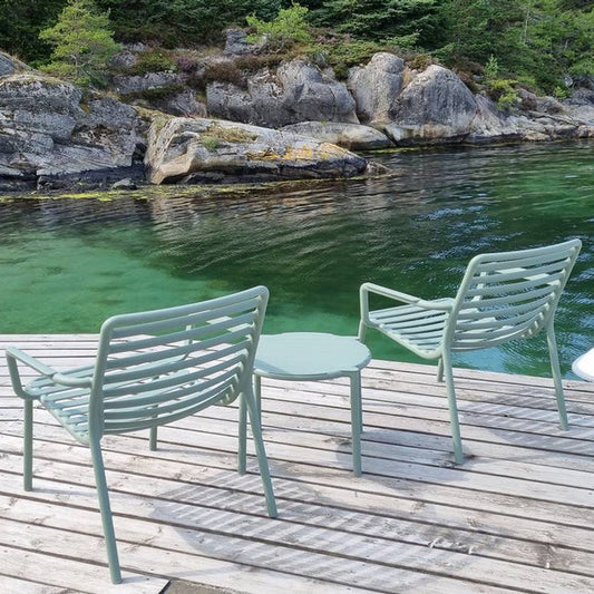 Three light sage green metal patio chairs and a matching small round side table arranged on a wooden dock overlooking clear turquoise water and rocky shoreline.