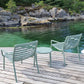 Three light sage green metal patio chairs and a matching small round side table arranged on a wooden dock overlooking clear turquoise water and rocky shoreline.