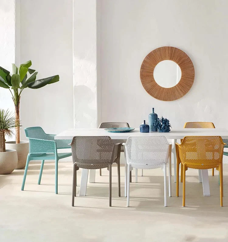 A dining table set with six modern mesh chairs in assorted colors including teal, gray, white, yellow, and brown, styled in a bright minimalist dining room with a round wooden wall mirror.