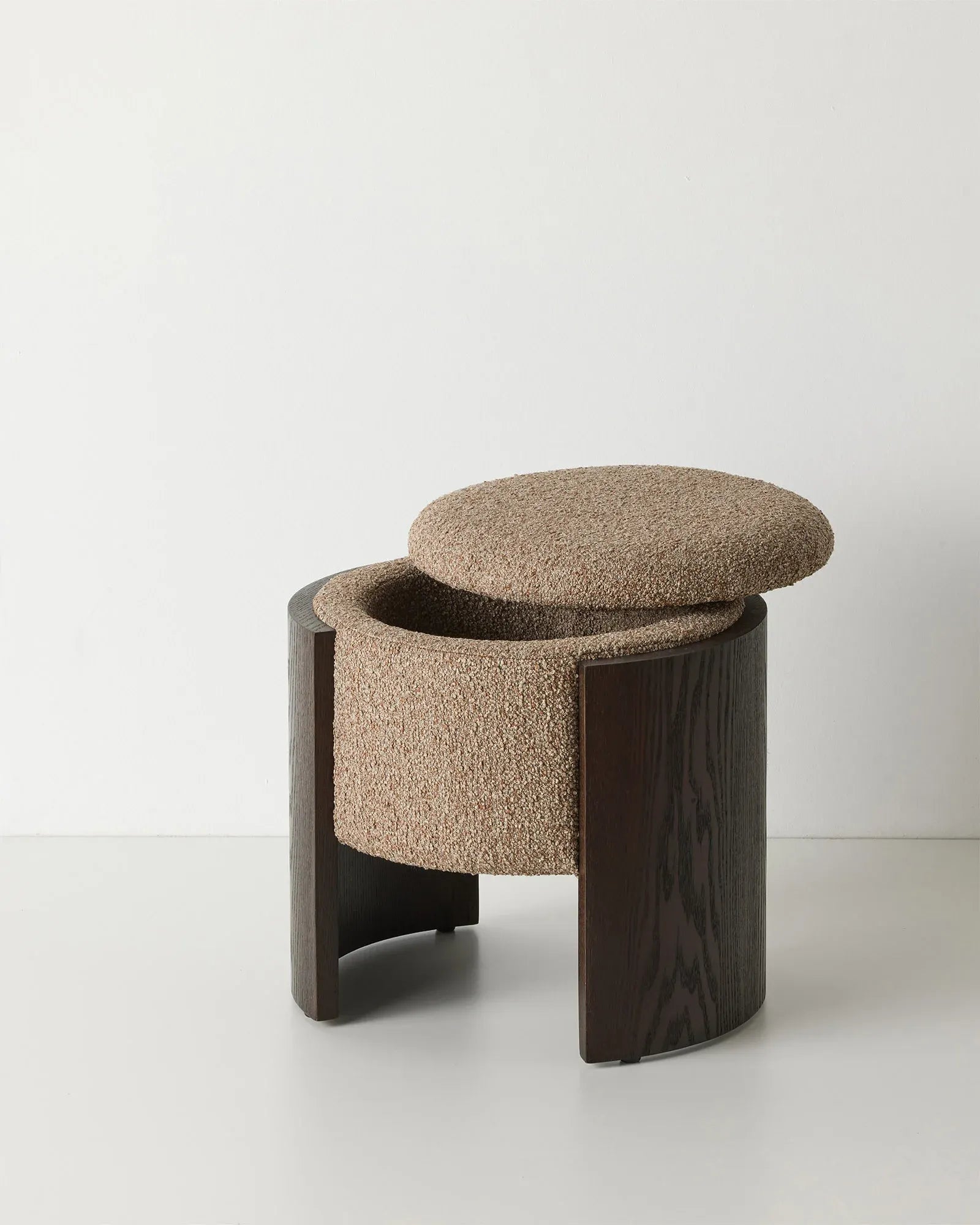 A modern low stool with a dark wood frame and a round tufted seat cushion upholstered in a textured beige fabric, set against a plain light gray background.
