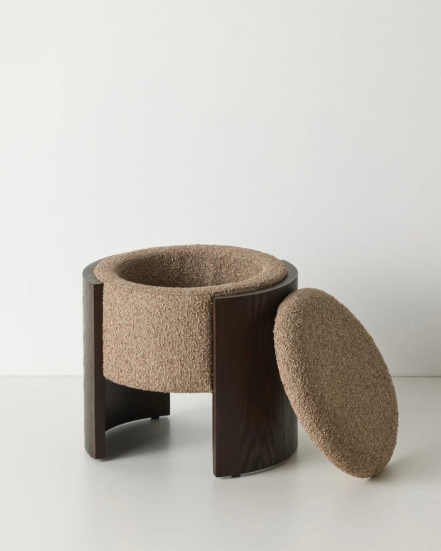 A modern cylindrical stool with a dark brown wooden frame and a textured beige bouclé fabric seat, shown with its matching oval cushion leaning against it on a light gray studio background.