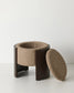 A modern cylindrical stool with a dark brown wooden frame and a textured beige bouclé fabric seat, shown with its matching oval cushion leaning against it on a light gray studio background.