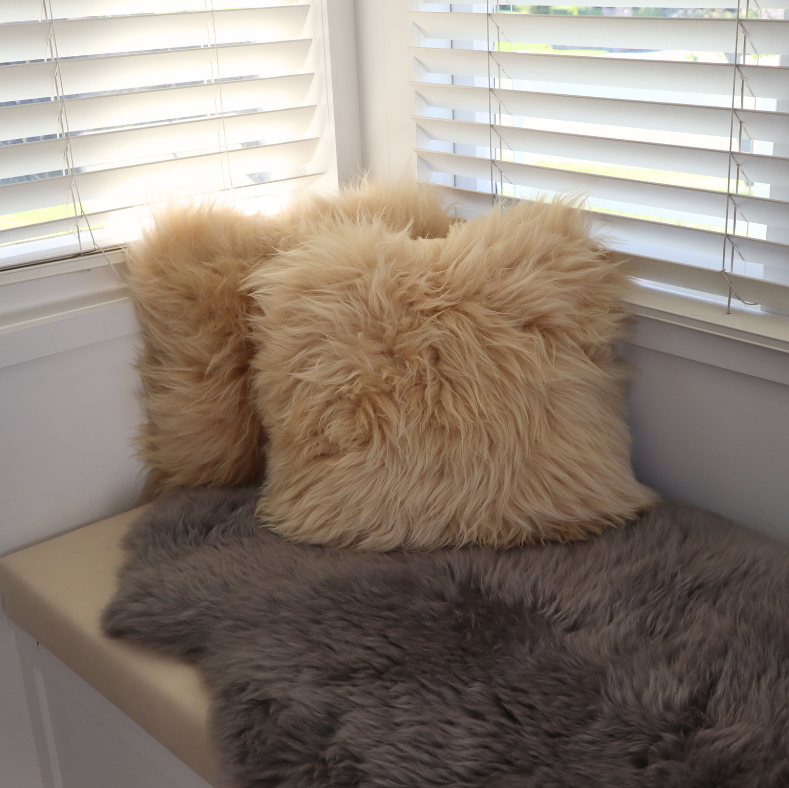 Two fluffy faux-shearling throw pillows in beige and cream, styled on a beige window seat with a gray faux-fur throw blanket in front of white window blinds.
