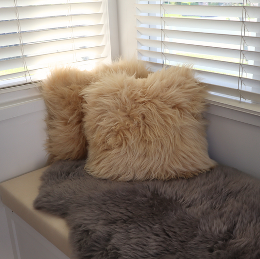 Two fluffy faux-shearling throw pillows in beige and cream, styled on a beige window seat with a gray faux-fur throw blanket in front of white window blinds.
