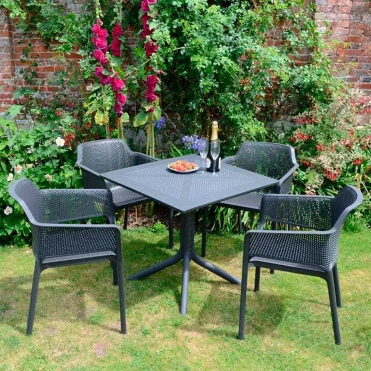 Outdoor garden setting with a black metal table and four chairs on a grassy area with flowers and a brick wall in the background.