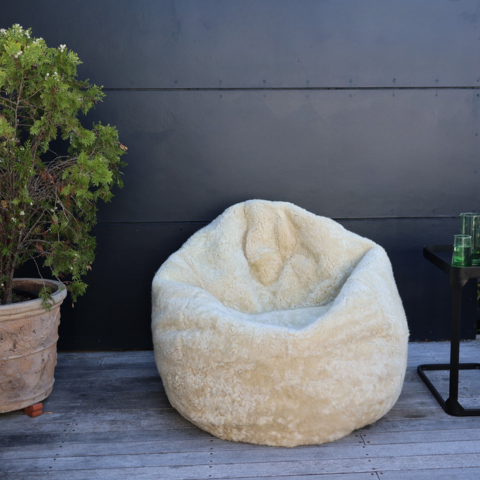 A plush, oversized bean bag chair upholstered in a textured cream or off-white sheepskin-like material, set on a gray wooden deck against a dark charcoal wall. A potted green plant is on the left, and a small black side table with green glasses is on the right.