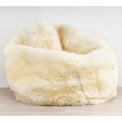A round, plush sheepskin or faux-sheepskin bean bag chair in a warm cream or ivory color, set against a neutral light gray wall and light wood floor.