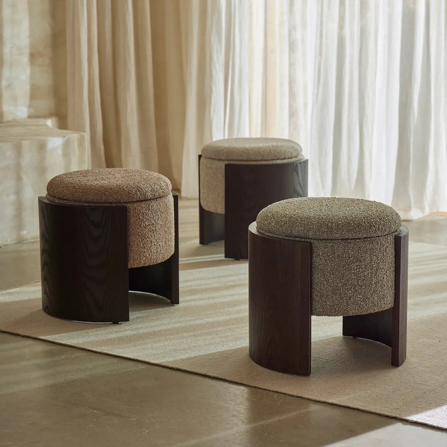 Three modern upholstered ottomans with dark wood frames arranged on a light rug in a softly lit room with sheer curtains.