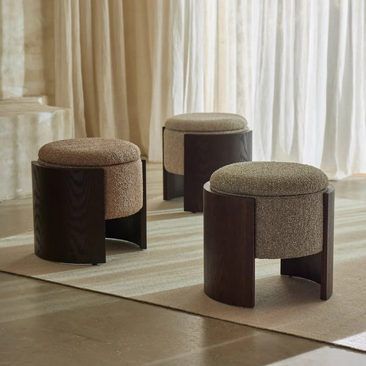 Three modern upholstered ottomans with dark wood frames arranged on a light rug in a softly lit room with sheer curtains.