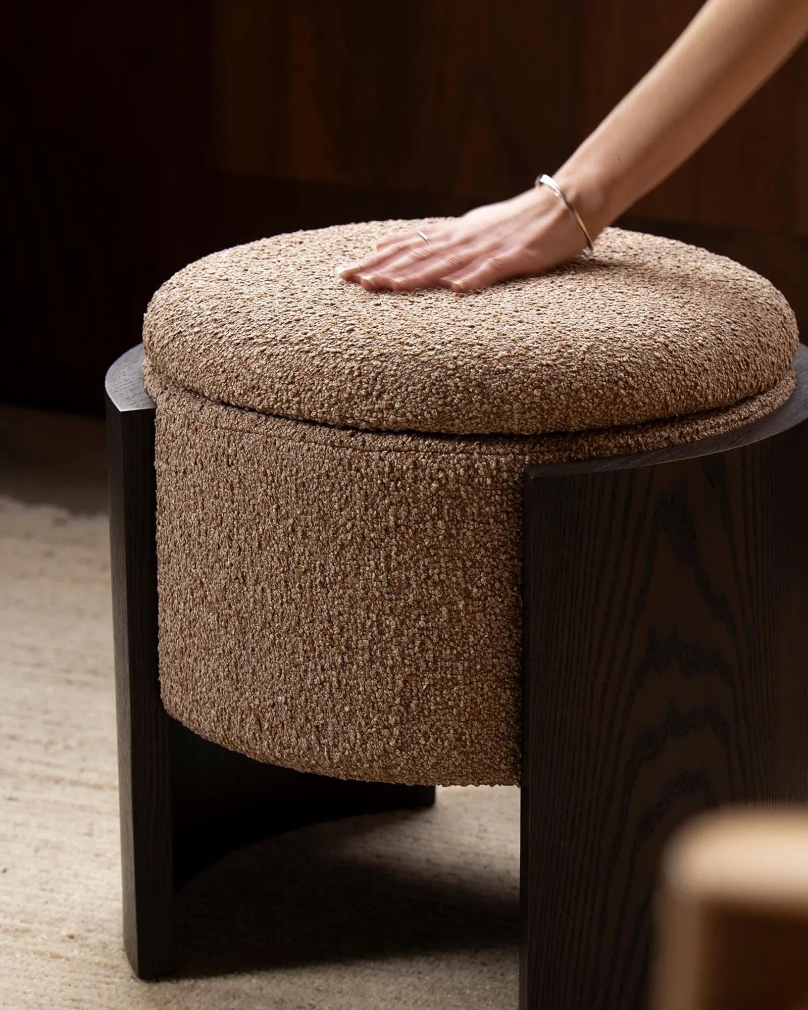 A round upholstered ottoman stool with a textured beige bouclé fabric cushion and dark wood legs, shown with a person's hand resting on the seat.