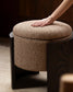A round upholstered ottoman stool with a textured beige bouclé fabric cushion and dark wood legs, shown with a person's hand resting on the seat.
